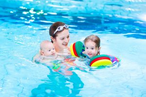 Young active mother playing with her toddler daughter and baby son in a swimmin